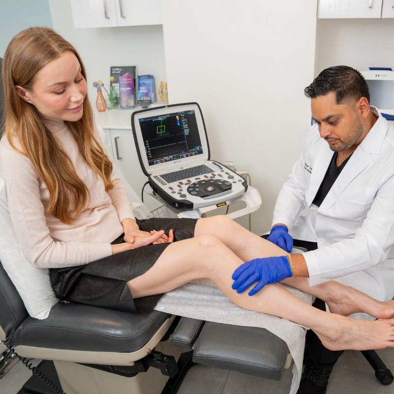 VTC_DrSadloo_Lifestyle_ (24)_7008x4672_optimized Dr. Ryan Sadloo is performing a physical examination on a female patient's leg in a clinical setting. He is using an ultrasound machine to conduct a diagnostic assessment of the patient's veins.